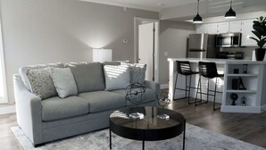A modern living room with a grey sofa, a round coffee table, and a kitchen area with white cabinets and stainless steel appliances. - One Bedroom,  First Floor (Wichita)