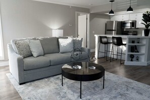 Ein modernes Wohnzimmer mit grauem Sofa, einem runden Couchtisch und einer Küchenzeile mit weißen Schränken und Edelstahlgeräten.