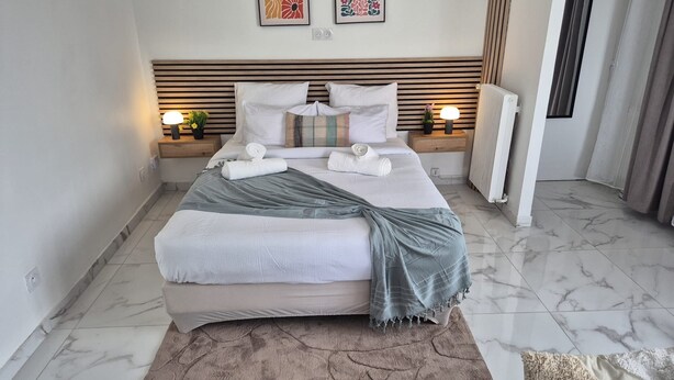A neatly made bed with a grey blanket, white pillows, and a striped throw. There are two bedside lamps, a radiator, and a marble floor. - Le Cocon : Bright studio 32m² + free private parking (rare). Near Stadium (Troyes)