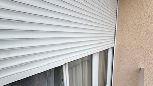 A window with white blinds, a clear view of the outside, and a small patch of artificial grass. - Le Cocon : Bright studio 32m² + free private parking (rare). Near Stadium (Troyes)