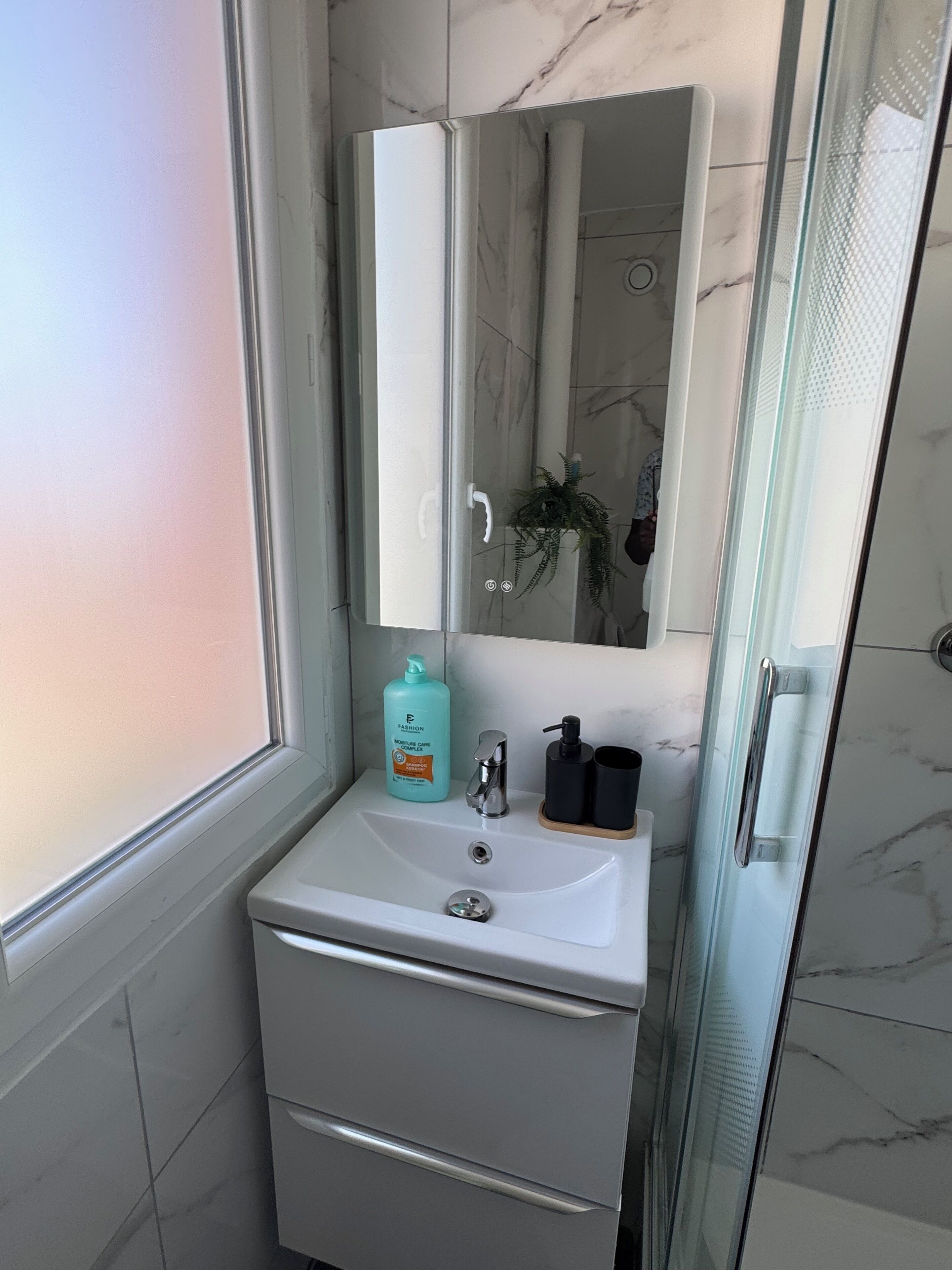 Een badkamer met een wand betegeld met marmer, een glazen douchecabine, een wit wastafelmeubel met kraan, een spiegel en een flesje vochtinbrengcrème.