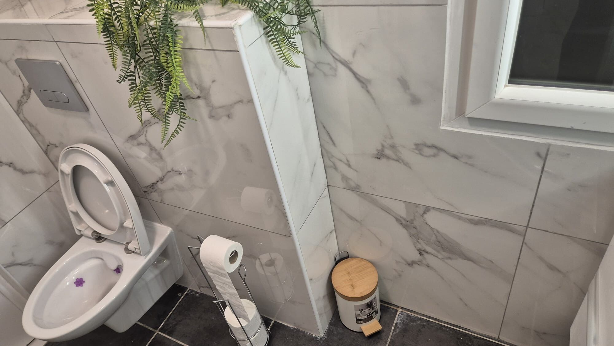 A bathroom with a toilet, marble walls, a plant, and a trash can.