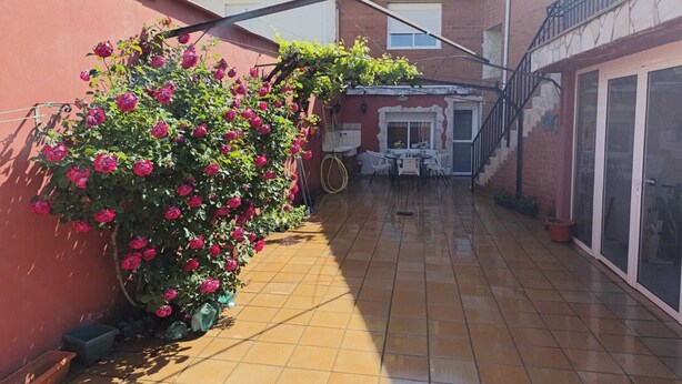 Un’area patio con pavimento piastrellato, un cespuglio di rose, un tavolo con sedie e un edificio con un muro rosso e una finestra.