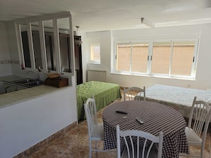 Una habitación con cocina, dos camas con colchas a cuadros, una mesa de comedor con sillas y una ventana con persianas.
