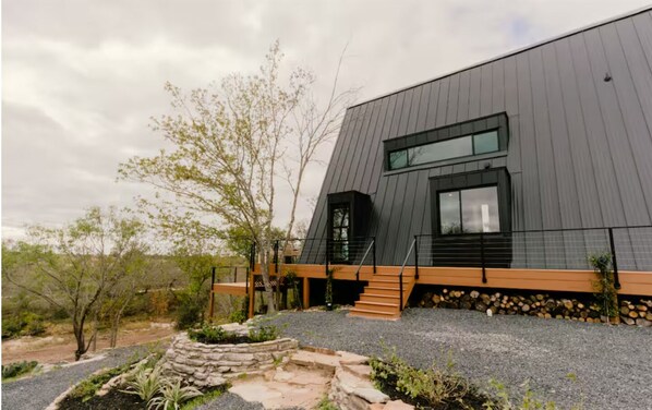 A modern building with a sloped roof, a wooden deck, and a stone pathway leading to the entrance. - NEW | The Pedernales A-Frame (Fredericksburg)