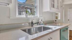 A kitchen with a stainless steel sink, a window with a view of a garden, and white cabinetry. - “Henry’s Haus” | Walk to Main | Pet Friendly! (Fredericksburg)
