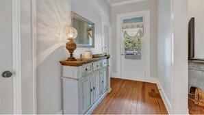 A hallway with a white door, a wooden floor, a white cabinet with a mirror, and a window with a view of the outside. - “Henry’s Haus” | Walk to Main | Pet Friendly! (Fredericksburg)