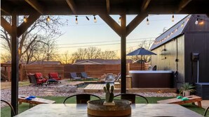 Een buitenpatio met een houten tafel, stoelen, een cactusschildering, een jacuzzi en een overdekte zithoek met lichtslingers.