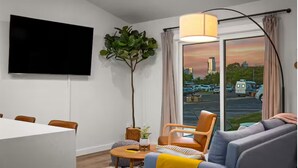 Una sala de estar moderna con televisión de pantalla plana, un ventanal con cortinas, un sofá, una silla y una mesa de comedor.