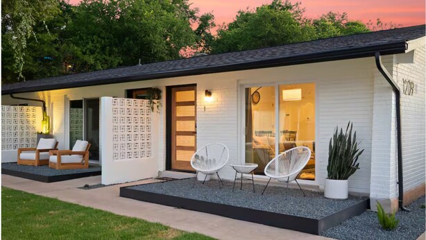 Un edificio moderno con techo plano, una puerta principal y un acceso de vidrio. Hay dos zonas de estar al aire libre con sillas y una pequeña mesa.