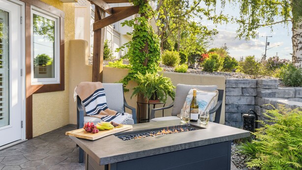 An outdoor patio with a stone fire feature, seating, a table with a bottle and glasses, and a potted plant. - Upper Mission Casita | Boutique Lakeview Suite for Business or Leisure (Kelowna)