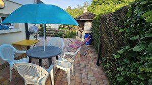 Een terras met een ronde tafel en vier stoelen, een blauwe parasol en een barbecue grill op de achtergrond.
