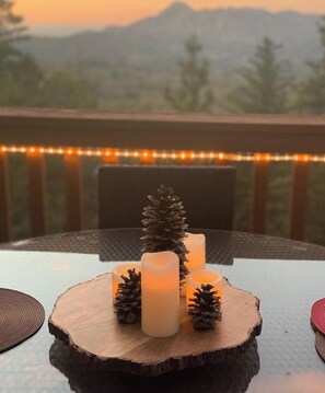 Ein Tisch mit Kerzen und Kiefernzapfen auf einer Holzplanke, platziert auf einem Balkon mit Bergblick und Lichterketten.