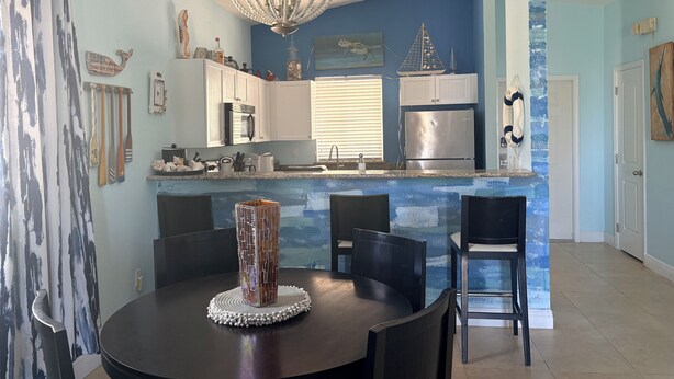 A dining area with a round table, chairs, a decorative centerpiece, a kitchen with white cabinets, a refrigerator, and a blue accent wall. - Fully Furnished 2BR/2BA Bimini Bay Condo (Bimini)