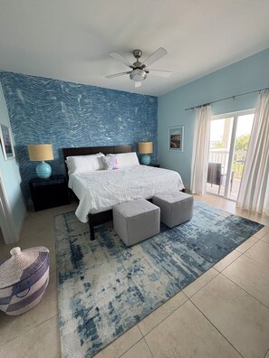 Une chambre à coucher comprenant un lit, deux tables de chevet, un ventilateur de plafond, une grande fenêtre avec des rideaux et un mur décoré d’un motif.