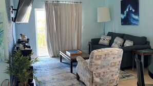 A living room with a ceiling fan, a sofa, a chair, a TV, a lamp, a painting, and a window with curtains. - Fully Furnished 2BR/2BA Bimini Bay Condo (Bimini)