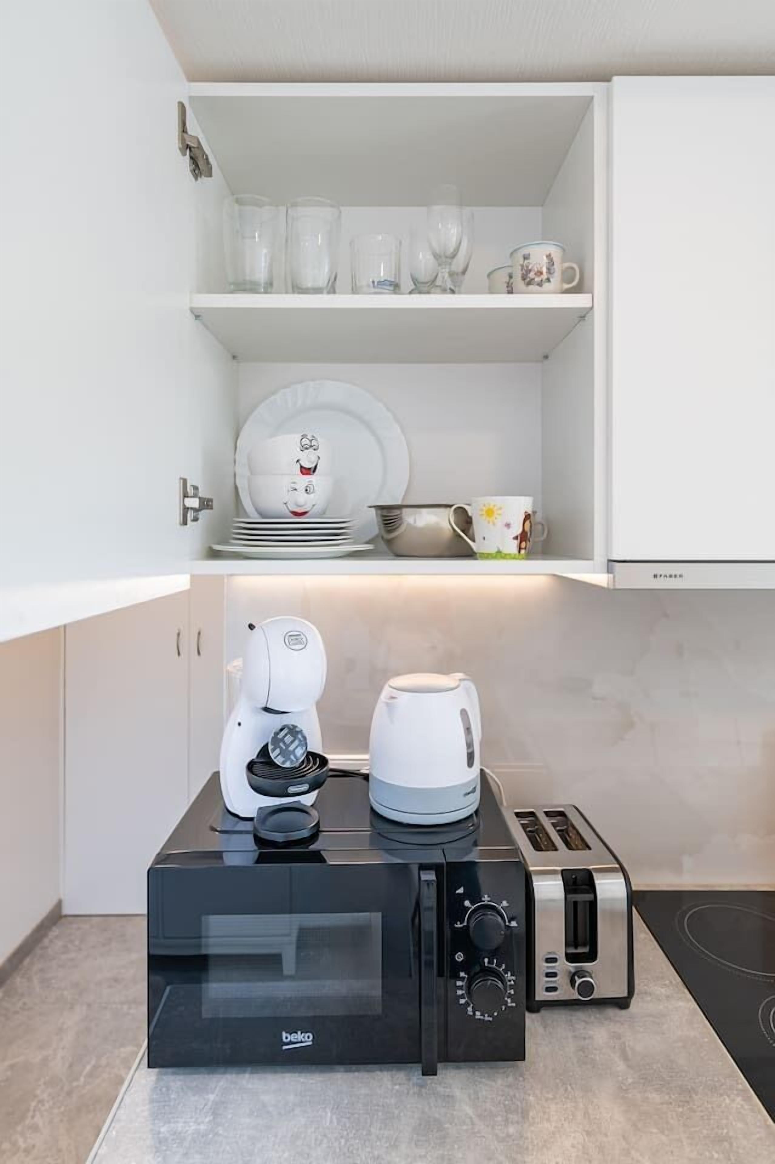 A kitchenette with a black microwave, a coffee maker, and a toaster. There are white shelves above containing glassware and plates.