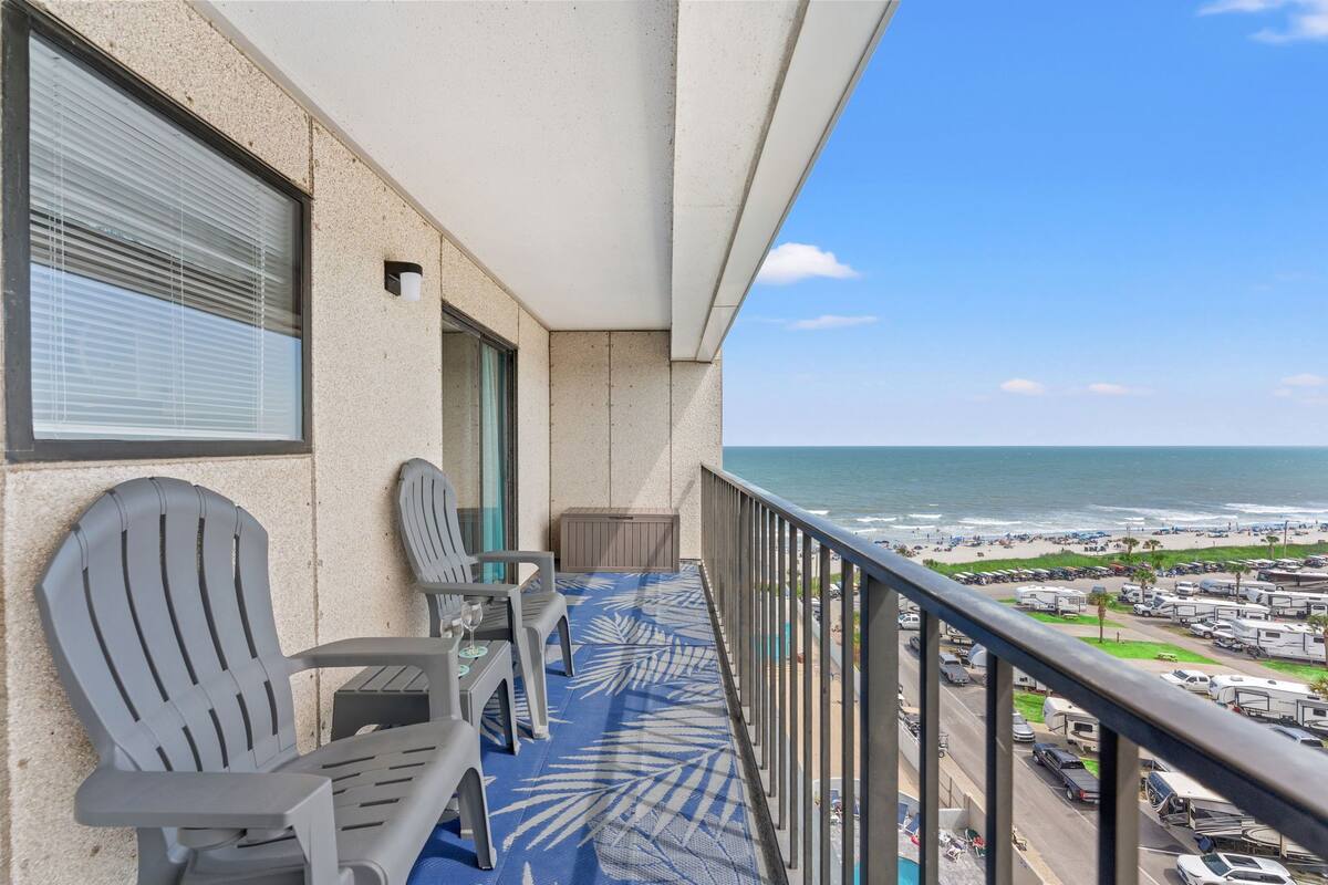 A balcony with two chairs, a table, and a view of the beach and ocean.
