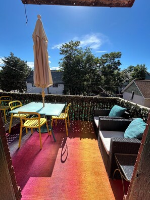 Eine Terrasse im Freien mit Tisch und Stühlen, ein Rattangeflechtsofa mit Kissen und einem Sonnenschirm.