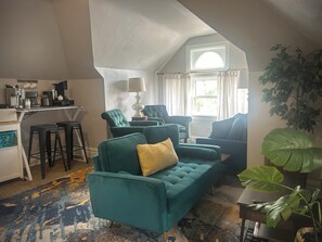 Ein gemütliches Wohnzimmer mit einem grünen Sofa, einer Kaffeestation und einem großen Fenster mit Vorhängen.