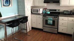 Una cucina con mobili bianchi, un piano cottura nero, un forno a microonde e un tostapane. C'è uno sgabello da bar e un quadro appeso al muro.