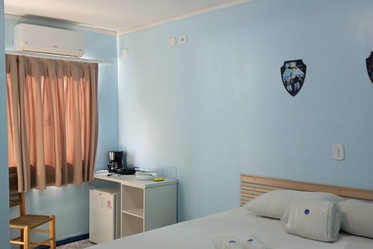 Habitación de hotel con cama, escritorio con cafetera, silla, equipo de aire acondicionado y lámpara fijada a la pared.