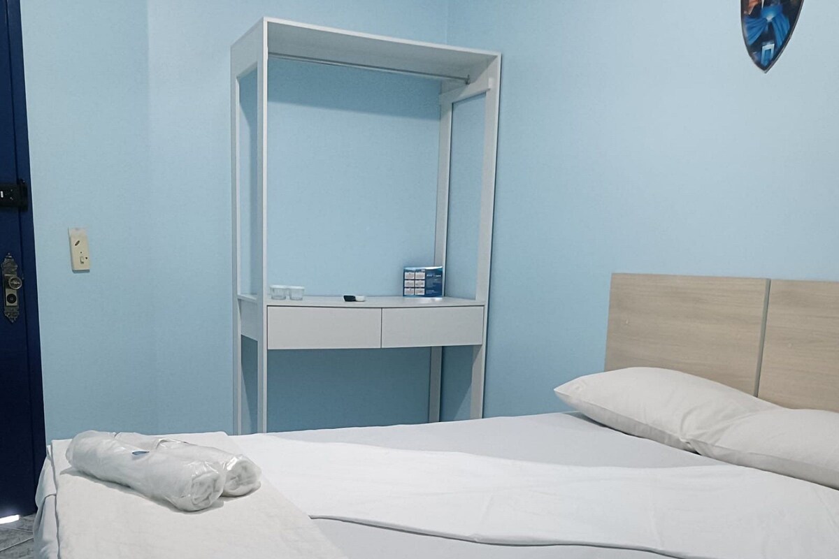 A neatly made bed with white linens, a wooden headboard, and a white bedside table with a mirror and a few items on top.