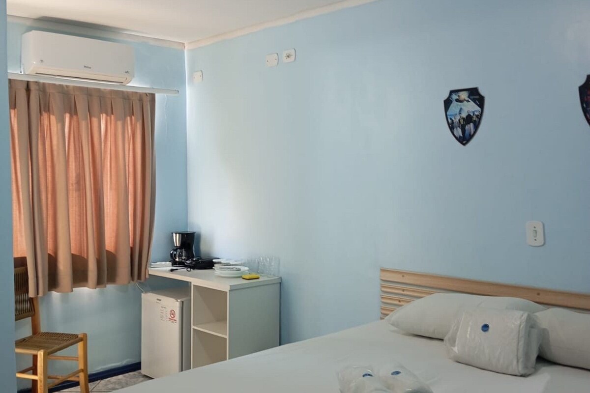 A hotel room with a bed, a desk with a coffee maker, a chair, an air conditioning unit, and a wall-mounted light fixture.