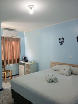 A hotel room with a bed, a desk with a coffee maker, a chair, an air conditioning unit, and a wall-mounted light fixture.