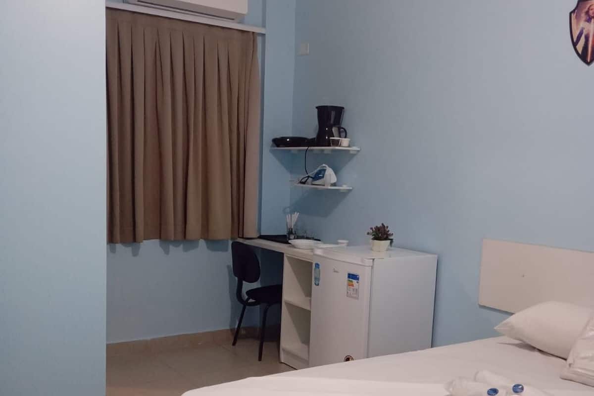 Una habitación de hotel con una cama, un escritorio, una silla, un refrigerador pequeño, una estantería con cafetera y un equipo de aire acondicionado.