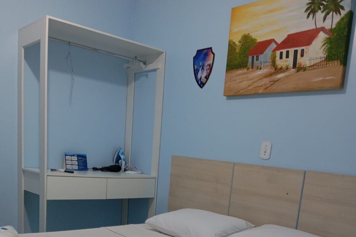 A bed with white linens, a wooden headboard, a small desk with a chair, a wall-mounted TV, and a painting of a house.