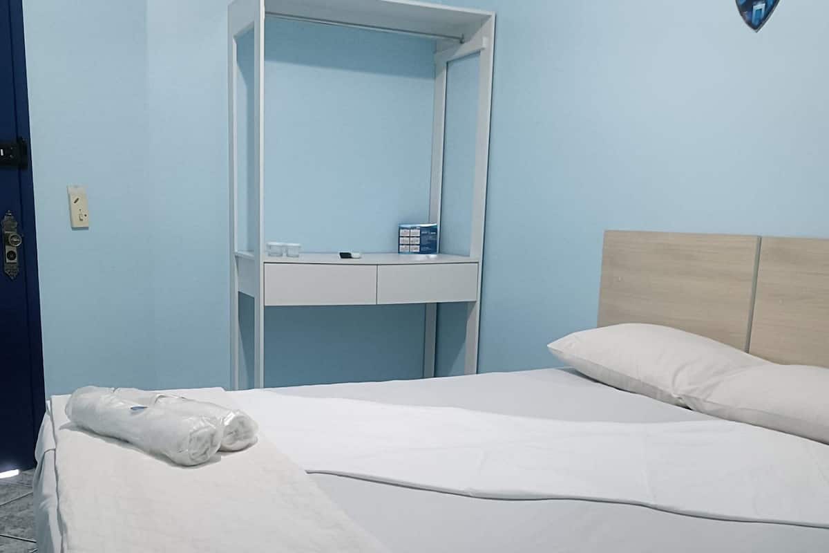 Una cama bien hecha con sábanas blancas, cabecera de madera y una mesita de noche blanca con un espejo y algunos objetos encima.