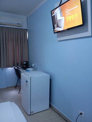 A hotel room with a bed, a desk with a coffee maker, a refrigerator, a TV mounted on the wall displaying an advertisement, and a window with curtains. - Residencial Wolverine  (Foz do Iguaçu)