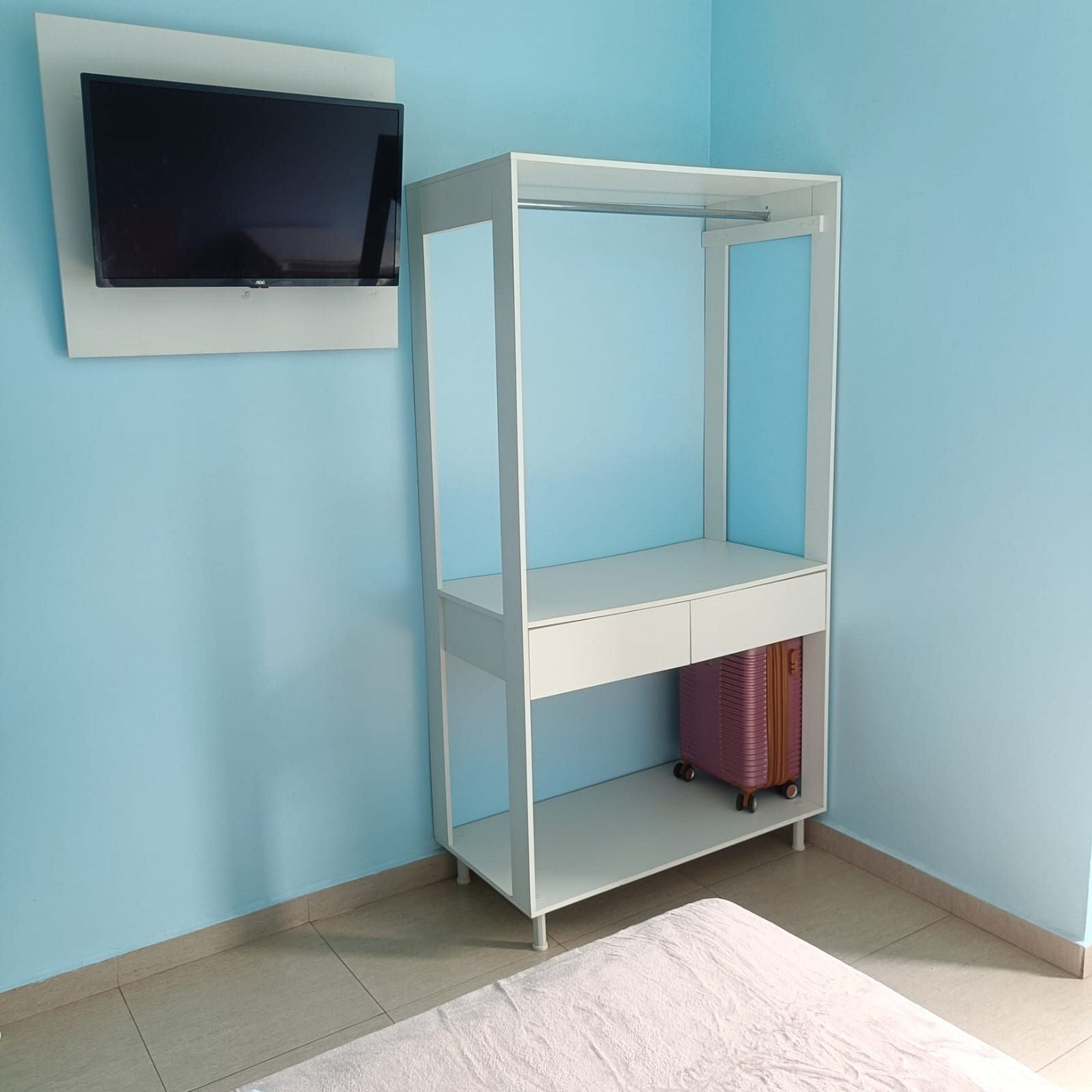 Ein Eckzimmer mit einem weißen Fernseher an der Wand, einer weißen Ablage mit einem pinken Koffer darunter und einem weißen Teppich auf dem Boden.
