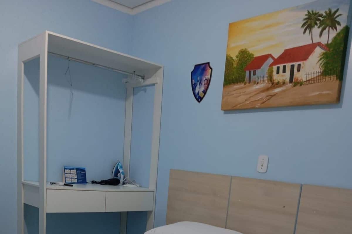 Una cama con ropa de cama blanca, un cabecero de madera, un escritorio pequeño con una silla, un televisor montado en la pared y un cuadro de una casa.
