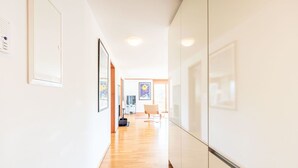 Un pasillo con suelo de madera, paredes blancas y cuadros enmarcados. Al final del pasillo hay una puerta que da a una habitación con una silla y un televisor.