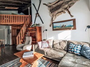 Ein Wohnzimmer mit einer Ledersofa, einem Holztisch, einem gemusterten Kissen, einem Gemälde und einem an Wand aufgehängten Tierfell.