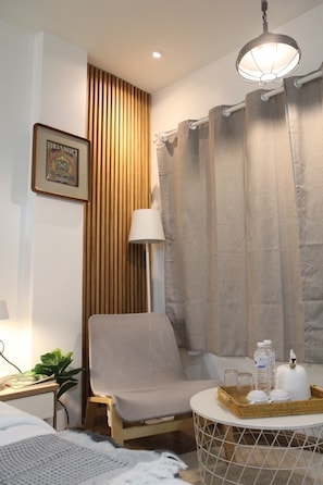 Ein Zimmer mit einem Sessel, einem kleinen Tisch mit Flaschen, einer Lampe, einem gerahmten Bild und einem Vorhang.