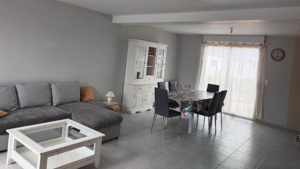 A living room with a sofa, a coffee table, a dining table with chairs, a cabinet, and a window with curtains. - New 3-bedroom bungalow (Mouzeil)