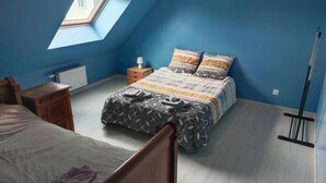 A bedroom with a sloped ceiling, a skylight, a bed with patterned bedding, a bedside table, a radiator, and a standing lamp. - New 3-bedroom bungalow (Mouzeil)