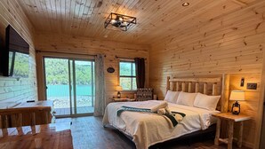 A wooden cabin bedroom with a bed, bedside tables, a desk, and a view through a sliding door. - Cedar - Big Moose Lakeside (Eagle Bay)