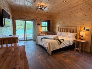 A wooden cabin bedroom with a bed, bedside tables, a desk, and a view through a sliding door.