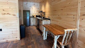 A compact kitchen with wooden walls, a stainless steel refrigerator, a wooden dining table, and a black trash bin. - Cedar - Big Moose Lakeside (Eagle Bay)
