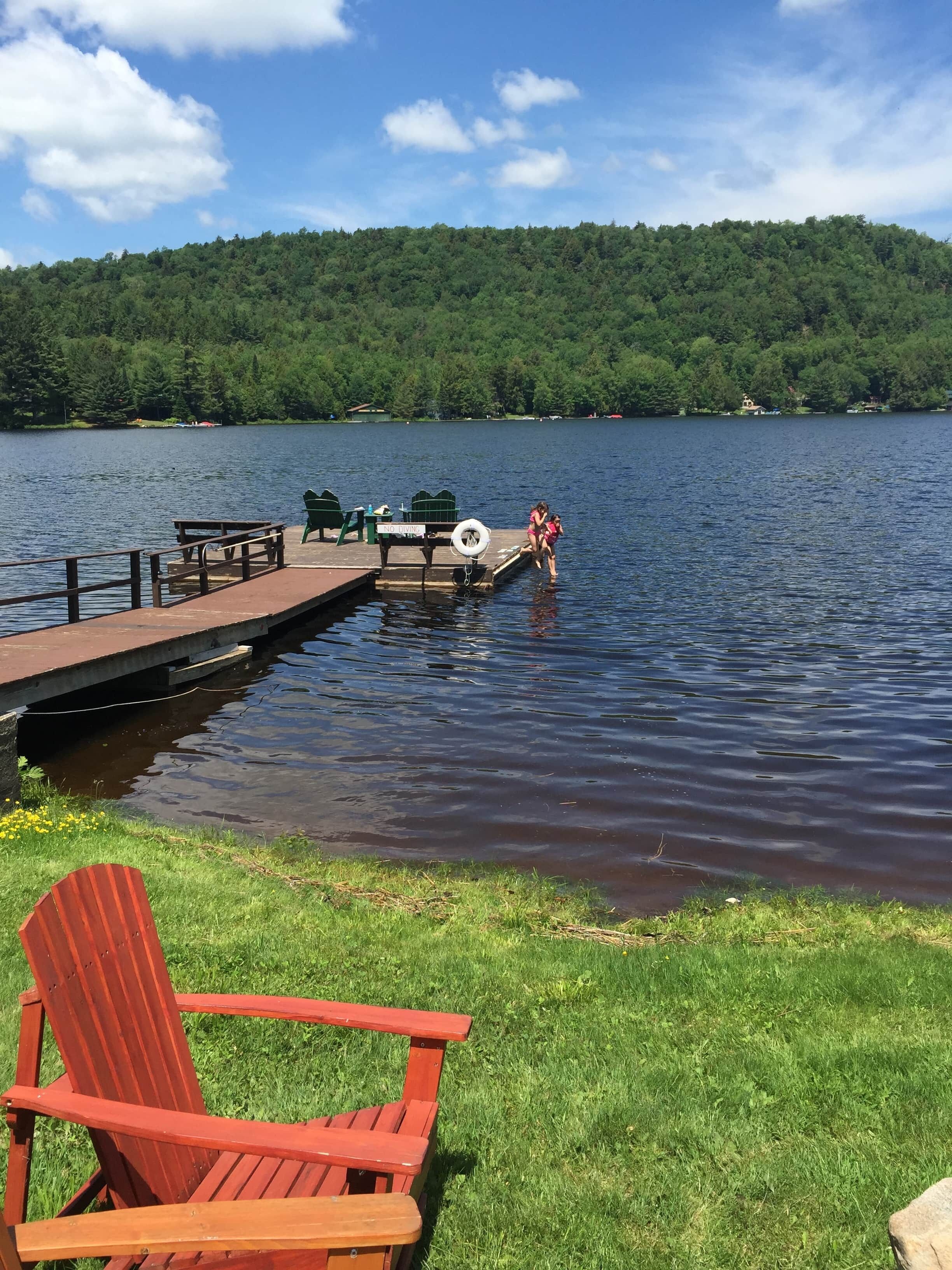 Um cais à beira do lago com uma cadeira Adirondack vermelha, uma placa com a inscrição 'NÃO MERGULHAR' e uma área gramada.