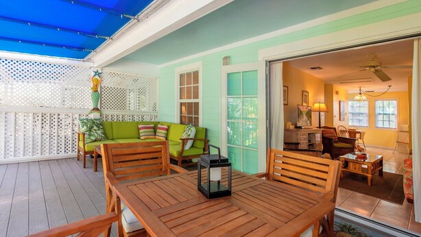 Eine Holzterrasse mit einem grünen Sofa und einem Tisch, ein weißes Gitter und der Blick in einen Raum mit Deckenventilator.