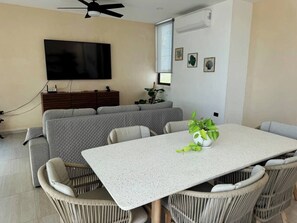 Ein modernes Wohnzimmer mit Flachbildfernseher, grauem Sofa, Esstisch, Korbstühlen und einer Topfpflanze.