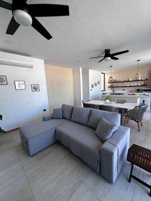 Ein modernes Wohnzimmer mit einem grauen Ecksofa, einem Essbereich mit rundem Tisch und einer Küche im Hintergrund.