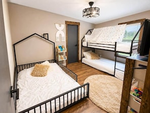 A bedroom with a bunk bed, a single bed, a wooden floor, a bookshelf, and a window with curtains.