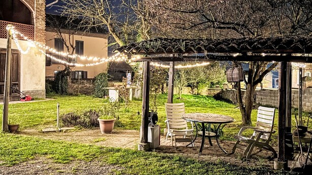 Un espace extérieur aménagé, abrité et doté d’une pergola, d’une table et de chaises, entouré d’un jardin et d’un bâtiment à la façade en pierre.