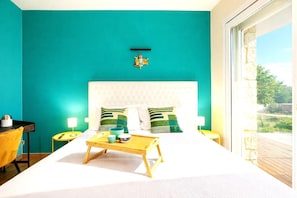 Ein Schlafzimmer mit einer türkisblauen Wand, einem weißen Bett und Blick ins Freie.